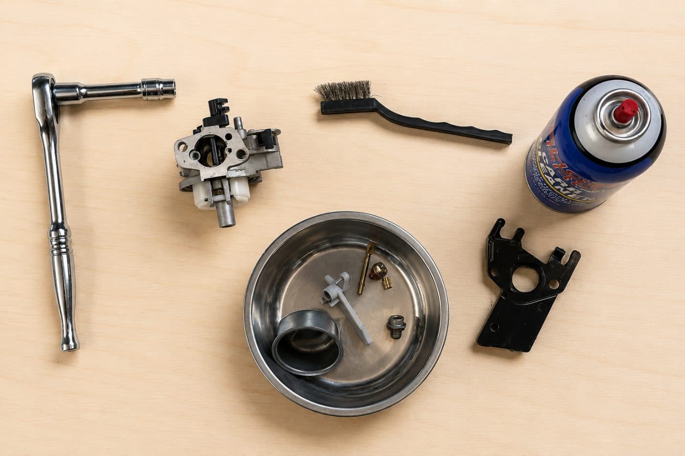 A ratchet, carburetor, wire brush and small parts laid out on a workbench