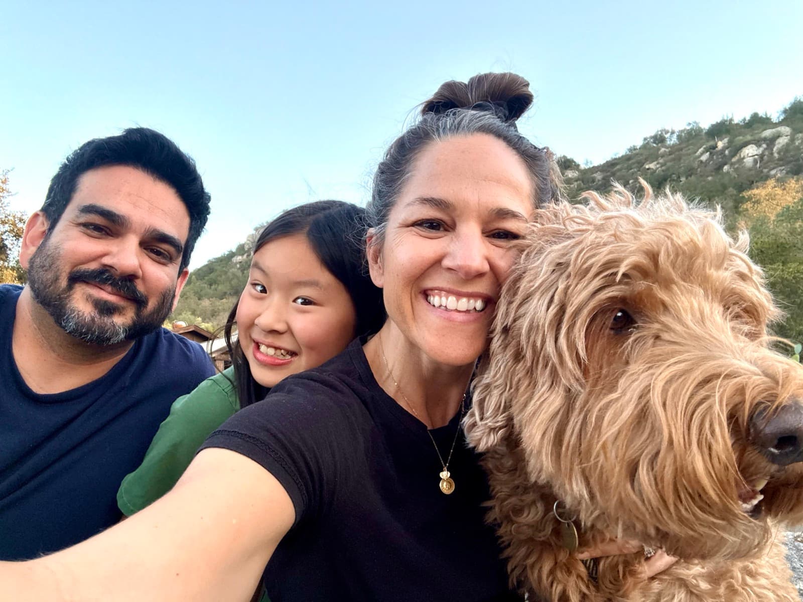 Becky, Jesse and their daughter smiling with their goldendoodle in front of the Valley Center foothills
